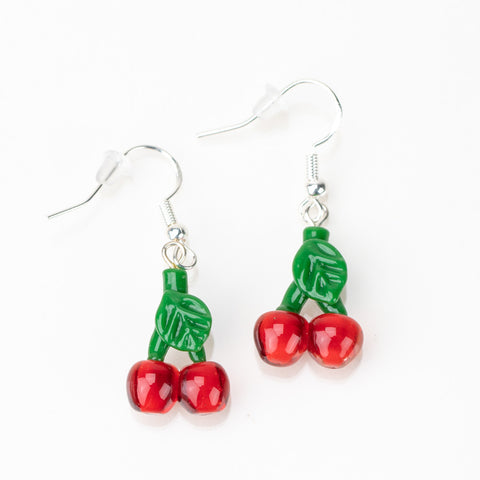 A set of cherry glass dangle earrings laying flat on a white table. The cherries are a bright red and feature a stem with a leaf design. The top of the earring has a sterling silver hook backing.
