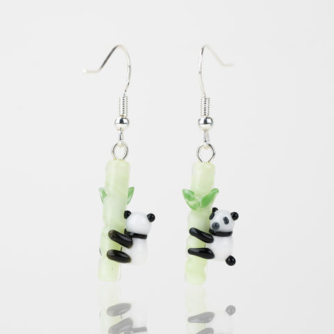 A couple of panda-themed dangle earrings with the left facing away from the camera and the right facing towards the camera. Each earring features a black and white panda hugging and green bamboo pole. Attached to the top of each bamboo pole is a sterling silver earring hook.