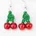 A close up of the "Cherries" glass dangle earrings from mmmelty. The cherries are a bright cherry red and the stem of the cherries is green with a leaf. The design is made completely of glass and feature a sterling silver earring backing.  