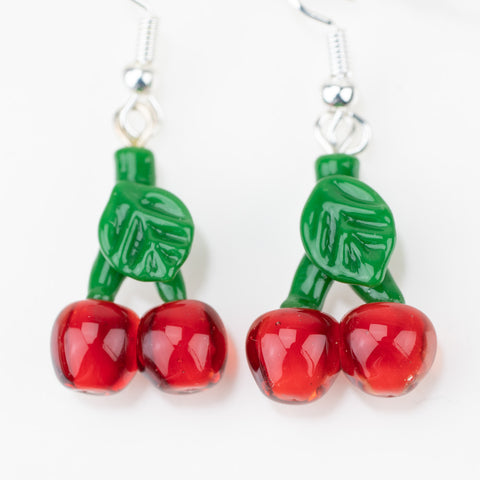 A close up of the "Cherries" glass dangle earrings from mmmelty. The cherries are a bright cherry red and the stem of the cherries is green with a leaf. The design is made completely of glass and feature a sterling silver earring backing.  