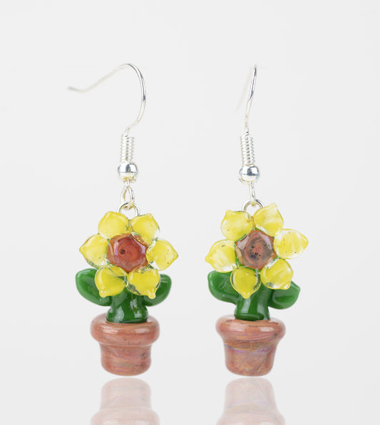 A pair of sunflower themed dangle earrings made of glass. The sunflower petals are yellow with a brown center, green stem and leafs, and a brown pot. Attached to each sunflower is a sterling silver earring backing. 