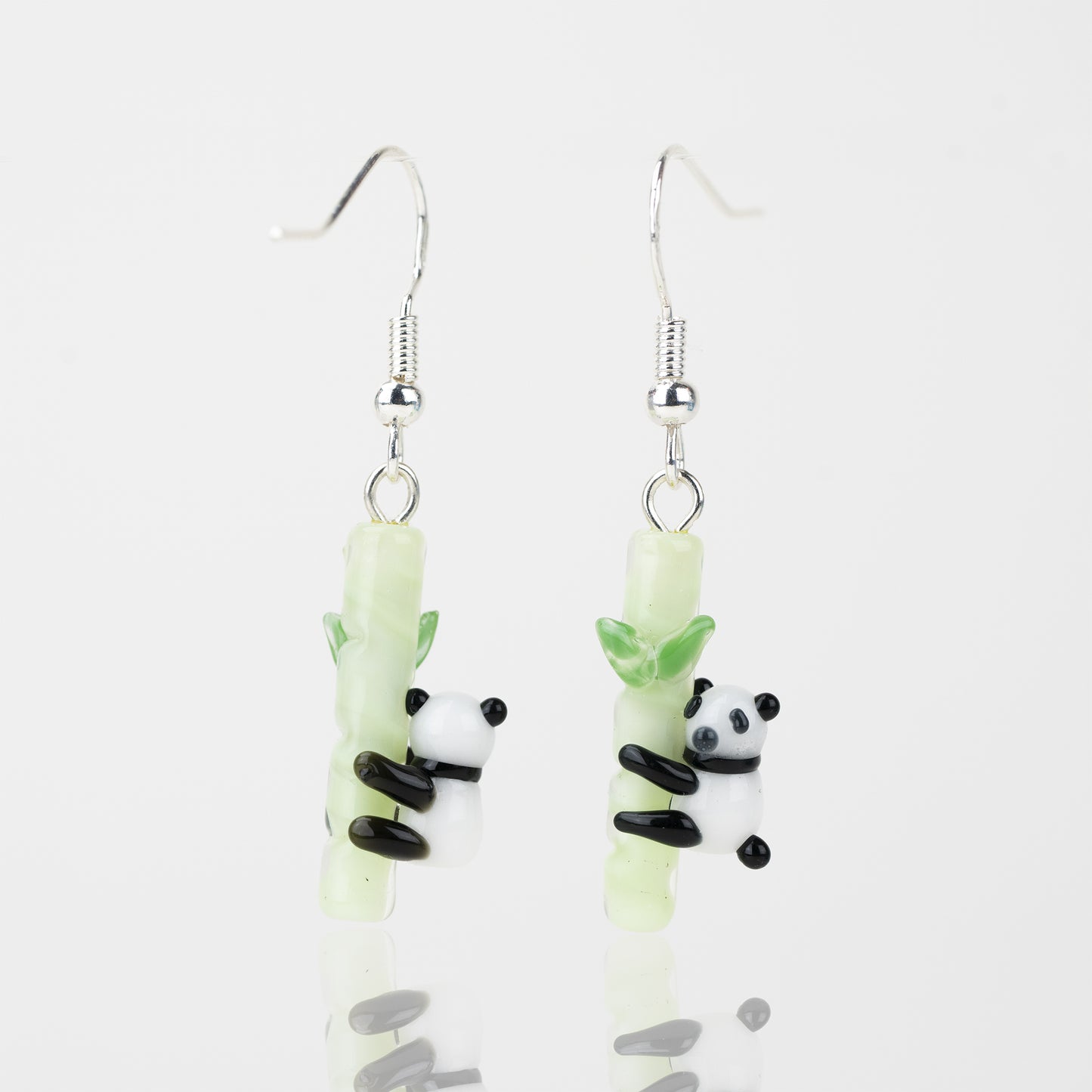 A couple of panda-themed dangle earrings with the left facing away from the camera and the right facing towards the camera. Each earring features a black and white panda hugging and green bamboo pole. Attached to the top of each bamboo pole is a sterling silver earring hook. 