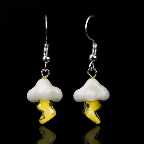 A couple of cloud and lightning dangle earrings on a black background. The cloud is white and the lightning bolt comes from the bottom of the cloud is yellow. Above each cloud is a sterling silver earring hook.