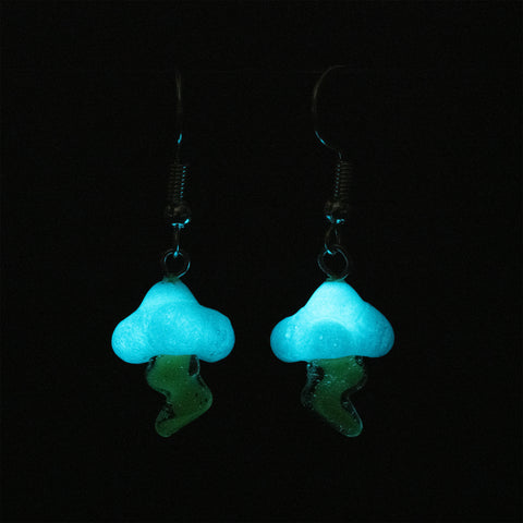 Two lightning cloud earrings under low light showcasing the glow in the dark features of the cloud. Below the cloud are the translucent lightning bolts.  